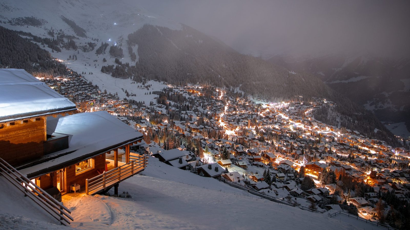 Chalet balcony above a valley of village lights in dusk snowfall